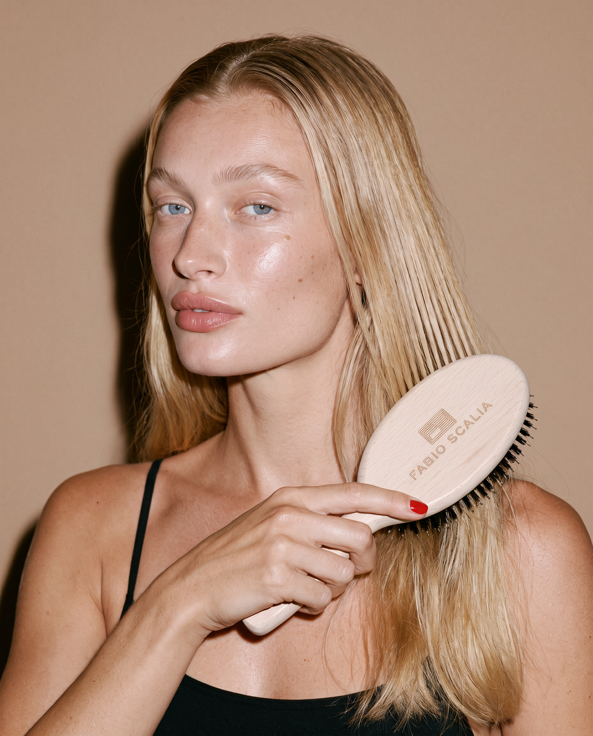 A blonde woman holds a light beige oval hairbrush with black bristles, branded 'FABIO SCALIA', against her hair, set against a solid warm beige background.